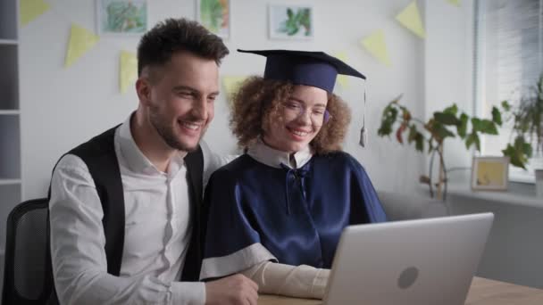 Jovem família alegre comemorar a graduação da faculdade e diploma com tecnologia de comunicação de vídeo moderna no laptop, enquanto sentado em casa — Vídeo de Stock