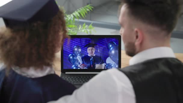 Távoktatás, női diák egyetemi öltözékben néz diplomaosztó ünnepség együtt a srác segítségével modern video kommunikációs technológia laptop — Stock videók