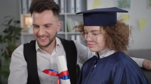 Выпускной, счастливая студентка в мантии и шляпе обнимает мужчину и радуется полученному сертификату об образовании онлайн по видеоконференции — стоковое видео