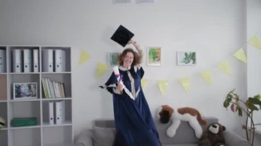 graduation celebrations, joyful female student in an academic mantle has fun jumping on couch with diploma in hands, takes off cap and throws it