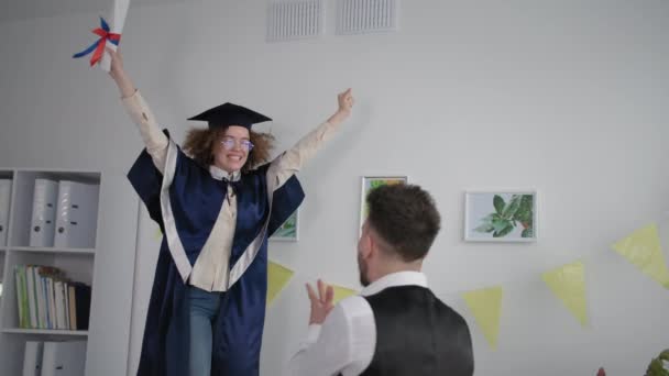 Girl in mantle and hat jumps on couch and hugs male friend rejoices at graduation from institute and certificate of education — Stockvideo