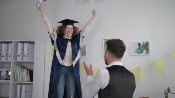 University graduation, cheerful student in academic clothes jumping on couch holding diploma and hugging male friend rejoicing at graduation — Stockvideo