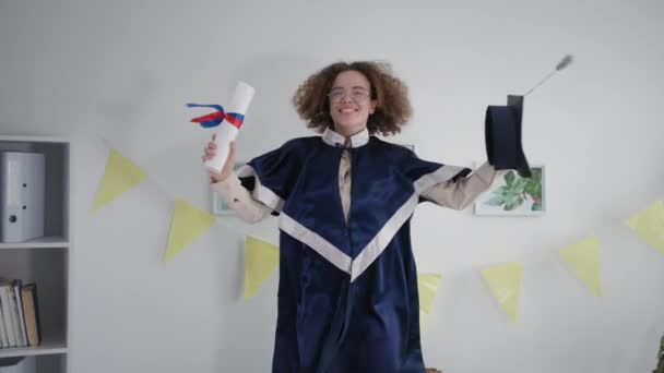 Female student in mantle huddles on couch with diploma and an academic cap in hands rejoicing at graduation from university — Αρχείο Βίντεο