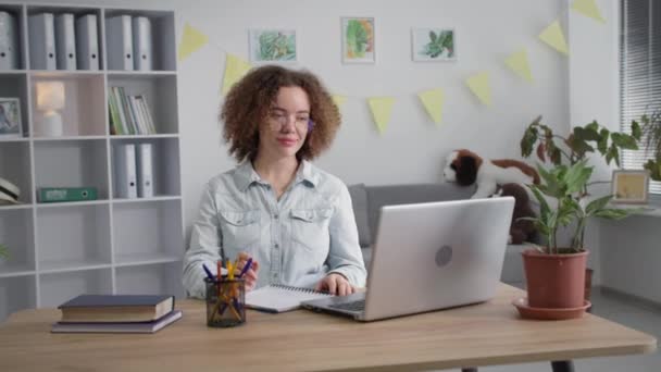 Menina adulta em óculos para visão ensina remotamente aulas com professor on-line via comunicação de vídeo no laptop escreve em notebook sorrindo e olhando para a câmera — Vídeo de Stock