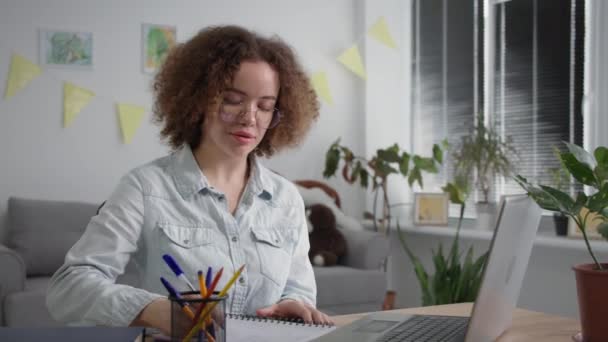 Обучение онлайн, молодая женщина в очках проходит дистанционное обучение разговаривая с учителем по видео связи на ноутбуке сидя за столом в комнате — стоковое видео
