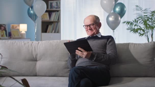 Grand-père s'amuse à envoyer des SMS sur les réseaux sociaux sur la tablette et à féliciter les vacances tout en étant assis sur le canapé fond de ballons dans le salon — Video