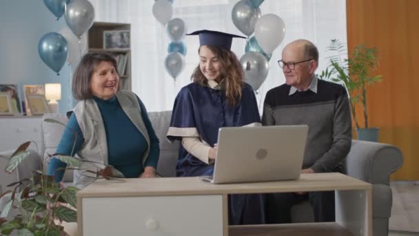 Щасливі батьки вітають випускницю та вручають їй диплом під час онлайн церемонії сидячи вдома у вітальні з повітряними кулями — стокове відео
