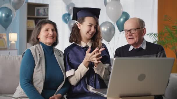 帽子の女の子とマントルは家に座っているラップトップ上の近代的な技術のウェブカメラの助けを借りてオンライン式中に卒業証書を提示お母さんとお父さんと喜び — ストック動画