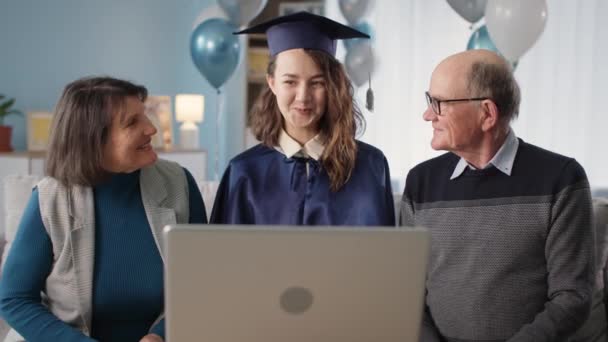 帽子とガウンを着た女性卒業生の家族は風船のリビングルームの背景に座っている間、ラップトップ上のビデオ通信を介してオンラインでお祝いを聞いています — ストック動画