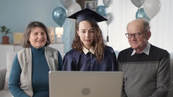 検疫でお祝いします,マントルと母親とお父さんと一緒に学術帽子を身に着けている若い女性学生は、ラップトップ上のビデオリンクを介して卒業証書をオンラインで受け取ります — ストック動画