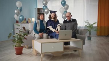 family with daughter in academic clothes celebrate online graduation ceremony while sitting at home on couch background of balloons about social distance time