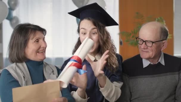 Menina alegre em chapéu acadêmico e vestido, juntamente com pais alegres celebra a formatura e graduação on-line — Vídeo de Stock