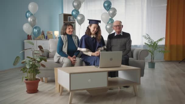 Modern education, student in a gown and an academic cap together with mom and dad celebrate online graduation ceremony while sitting at home on couch — Stok video
