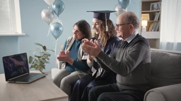 Glad universitet kandidat med forældre lykkeligt klappe deres hænder under graduering ceremoni online sidder hjemme i stuen – Stock-video