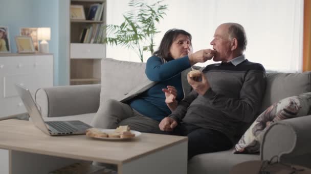 Szerető idős pár élvezze az időt együtt, és enni szendvics, miközben ül a kanapén a nappaliban — Stock videók