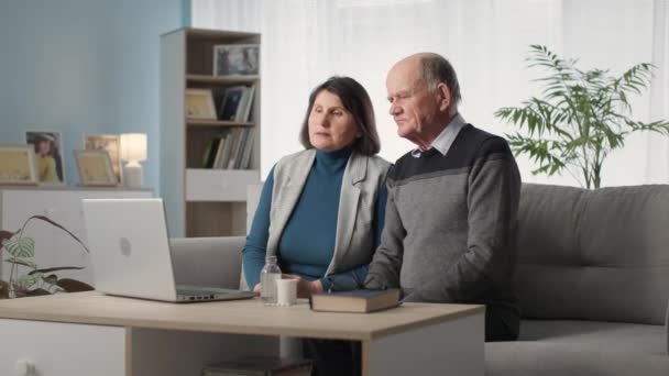 Подружня пара погано спілкується з лікарем онлайн через відеодзвінок на ноутбук, сидячи на дивані вдома — стокове відео