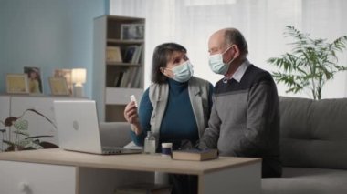 modern elderly man and woman in medical masks communicate by video call with family doctor about their health while sitting at home in living room
