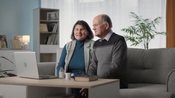 Ältere Frauen und Männer mit schlechtem Gesundheitszustand werden per Videolink auf dem Laptop mit dem Arzt über die Medikamentenwahl beraten, während sie im Wohnzimmer auf dem Sofa sitzen — Stockvideo