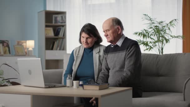 Літня пара розмовляє з медичним працівником через відеозв'язок на ноутбуці, сидячи вдома у вітальні — стокове відео