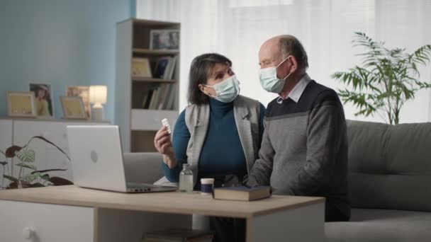 Modern elderly man and woman in medical masks communicate by video call with family doctor about their health while sitting at home in living room — Wideo stockowe