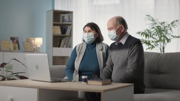 Hombre y mujer en máscaras médicas protección contra el virus y la infección comunicarse con el médico asistente en línea a través de la comunicación de vídeo en el ordenador portátil sentado en el sofá en la habitación — Vídeos de Stock
