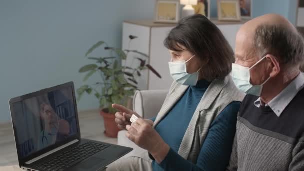 Пожилые муж и жена в масках на лицах общаться с врачом онлайн с помощью современных технологий видеосвязи на ноутбуке, сидя дома на диване во время пандемии — стоковое видео