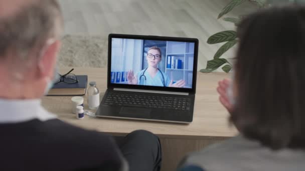 Пожилая пара в медицинских масках общаются с врачом онлайн, используя современные видеокоммуникации на ноутбуке, сидя дома во время карантина — стоковое видео