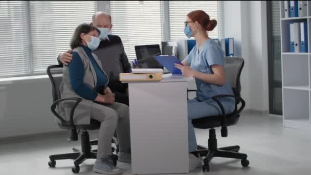 Doctor in medical clothes and face mask talks to an elderly man and woman about state of health in hospital office — Αρχείο Βίντεο