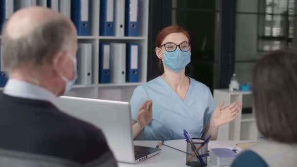 女医生戴着医疗面罩，在医院接待病人时与老年妇女及其丈夫就病人的健康和治疗进行交流 — 图库视频影像