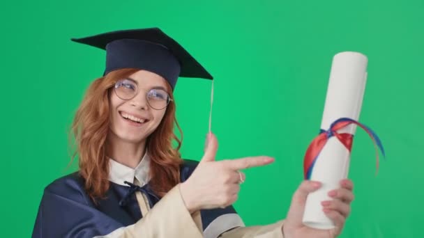 Young woman with glasses for vision is having fun and rejoices at graduation, holding diploma in hands and showing thumb up, green background — Stock Video