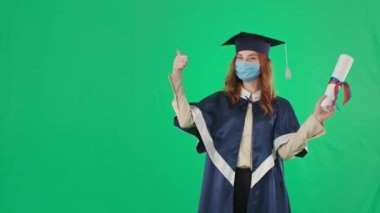 Mezuniyet mesafesi, önlüklü ve şapkalı genç bir kadın diploma almaktan memnun ve yeşil arka planda, krom anahtarda dikilirken başparmağıyla sınıfı gösteriyor.