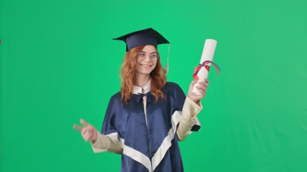 Graduación, mujer joven en una gorra académica y manto con diploma en las manos se regocija al final de los estudios de pie sobre fondo verde, croma clave — Vídeos de Stock