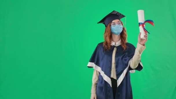 Kvinna examen i akademisk hatt och mantel i medicinsk mask på ansikte pekar på diplom, sedan finger upp och hand till sida plats för inskription när du står på grön bakgrund, kromnyckel — Stockvideo