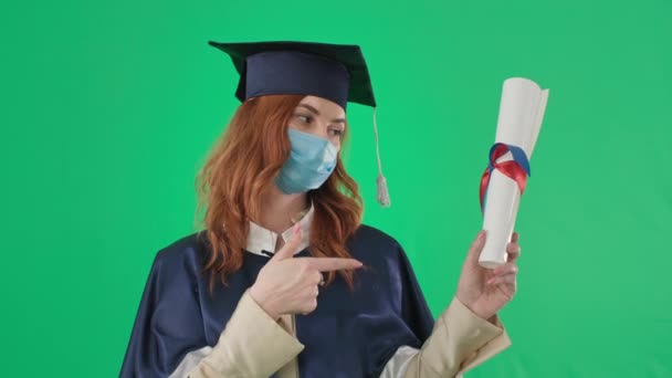Adult female student in medical mask and an academic gown and hat with diploma spreads arms to side on green background — Stock Video