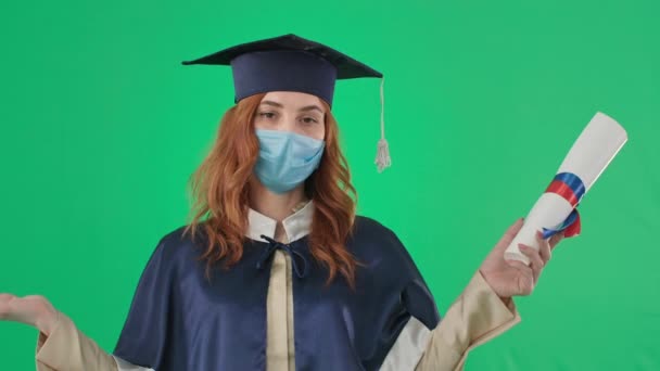 , donna laureata in abito accademico e maschera medica in possesso di diploma universitario punta il dito verso il basso, mentre lo studio on-line, chroma key — Video Stock