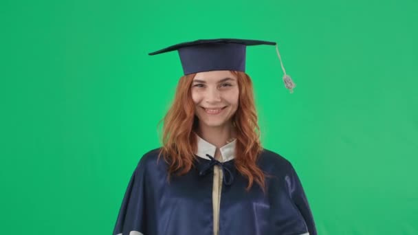 Graduation online, girl in academic clothes with diploma smiling and showing hand to class and camera on green screen background, chroma key — Stock Video