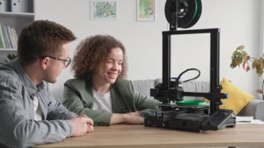 Modern teknolojiler, gülümseyen kadın ve erkek 3D modellemeyi tartışıyor ve evde otururken 3D yazıcı izliyor