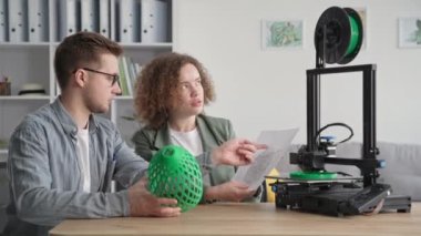 Yaratıcı erkek ve kadın modern teknolojiyi kullanır ve ellerinde 3D bir model tutarken 3D yazıcı evde otururken bir prototip basar.