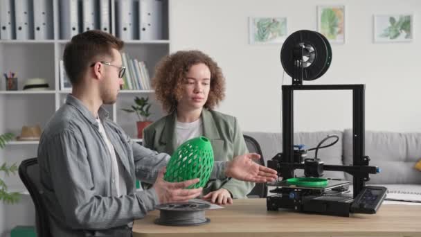 Mladá okouzlující žena a muž studují 3D model vyrobený na 3D tiskárně doma, zatímco sedí u stolu v pokoji — Stock video