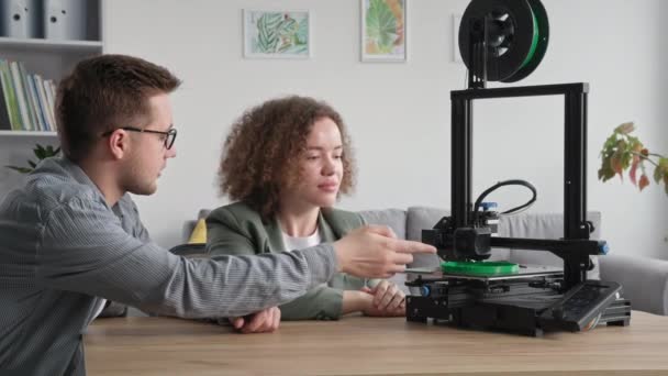 Młoda kreatywna para korzysta z nowoczesnej technologii i chętnie ogląda wydruk modelu 3D na drukarce 3D siedząc przy stole w domu — Wideo stockowe