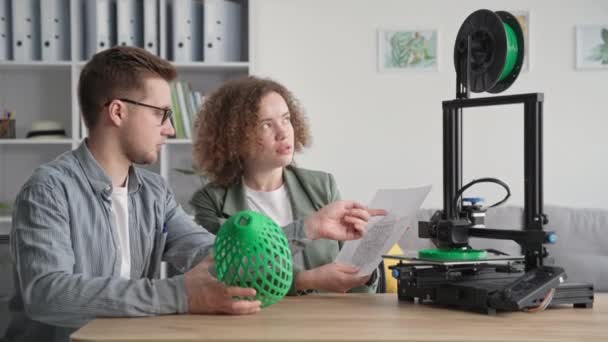 Hombre y mujer creativos utilizan la tecnología moderna y sostienen un modelo 3D en sus manos, mientras que una impresora 3D imprime un prototipo mientras está sentado en casa — Vídeos de Stock