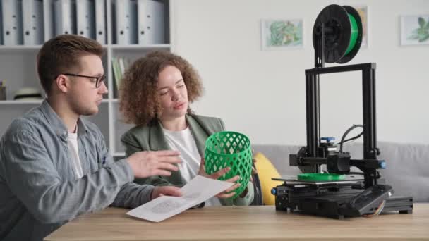 Moderne mannelijke en vrouwelijke ontwerpers kijken naar prototype print op 3D-printer en houden 3D-model in hun handen terwijl ze aan tafel zitten in de kamer — Stockvideo