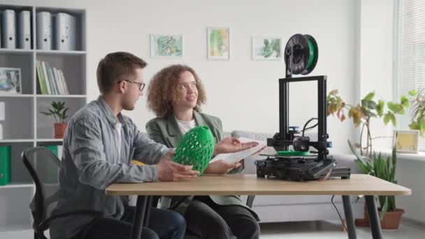 Innovative Technologien, glückliche männliche und weibliche Freunde verwenden 3D-Drucker zu Hause und sehen Prototypen drucken, während sie am Tisch im Raum sitzen — Stockvideo