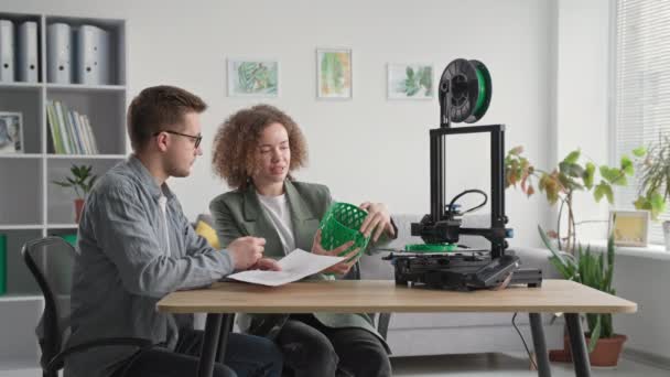 Современные инженеры женщины и мужчины используют современные технологии и обсуждают 3D модель в руке во время работы над 3D принтером, сидя за столом в комнате — стоковое видео