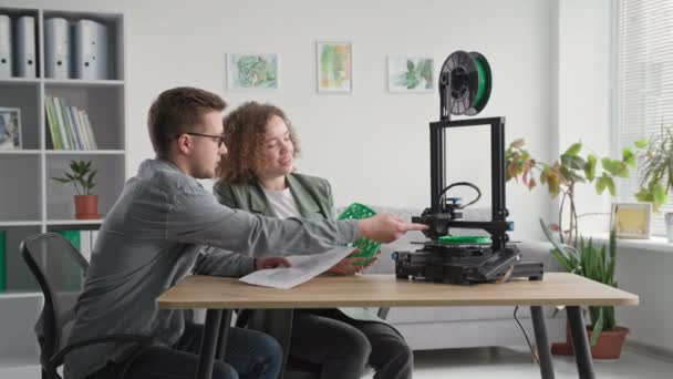 Ritratto di donna e uomo moderni che utilizzano la stampante 3D a casa e tengono in mano il modello 3D mentre si siedono a tavola nella stanza, sorridendo e guardando la fotocamera — Video Stock