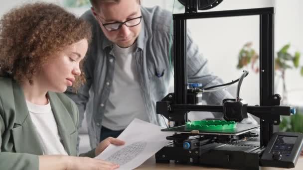 一个微笑的女人和一个男人一起使用现代技术，开心地坐在桌旁看3D打印机 — 图库视频影像