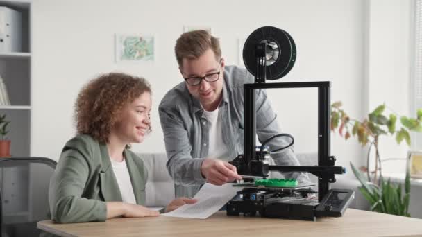 3D printing models at home, smiling girl with young guy are watching project for 3D printer sitting at table in room — Stock Video