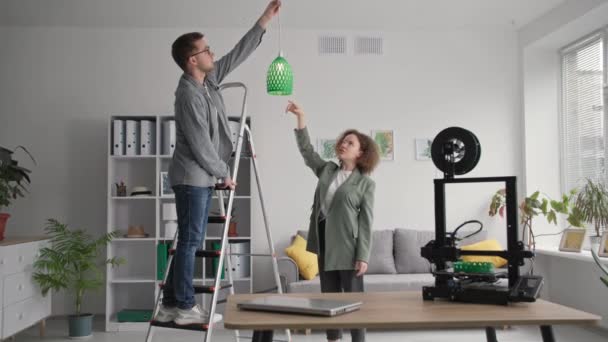 Portrait homme et femme créatifs font amélioration de la maison et accrocher l'ombre de lampe prototype imprimé sur imprimante 3D dans le salon, souriant et regardant dans la caméra — Video