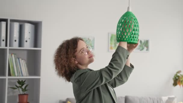 Portret van creatieve jonge vrouw met bril hangt bedrukte lampenkap op 3D-printer, glimlacht en kijkt naar de camera — Stockvideo