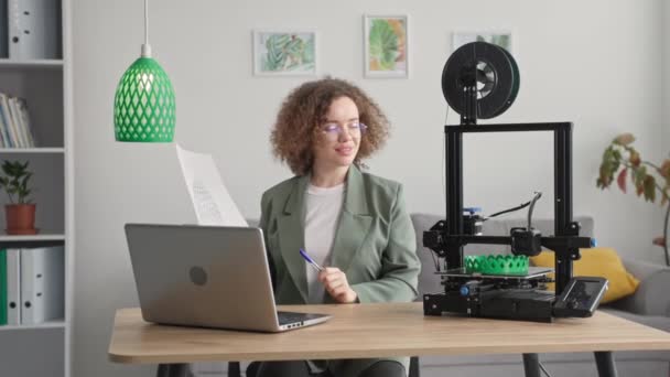 Portret van slimme creatieve vrouwelijke ingenieur maken prototype van 3d model op de computer van schaduw voor lamp tijdens het afdrukken op 3d printer terwijl zitten aan tafel in de kamer — Stockvideo